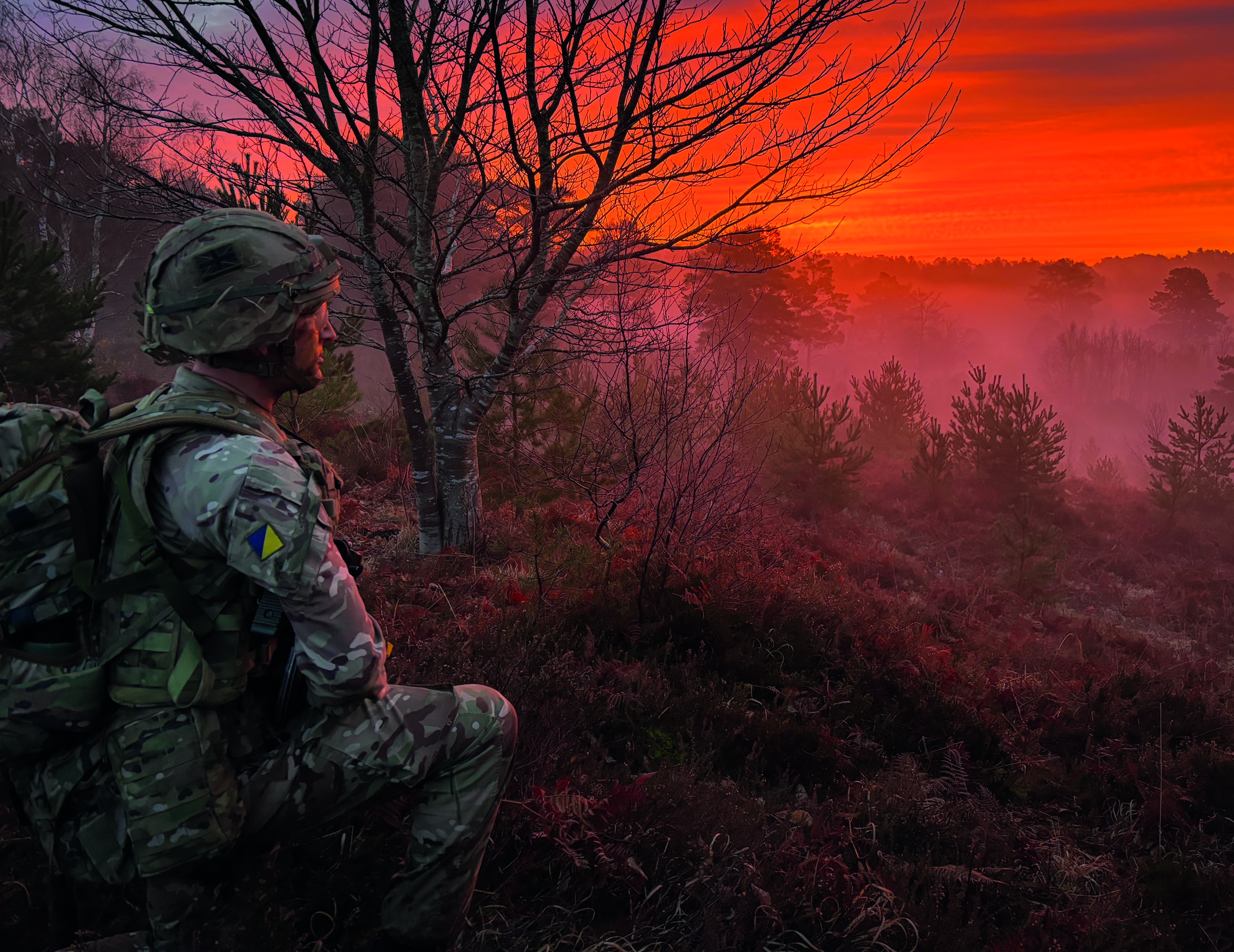 Uniformed soldier crouching and staring into a red sunset over wooded area