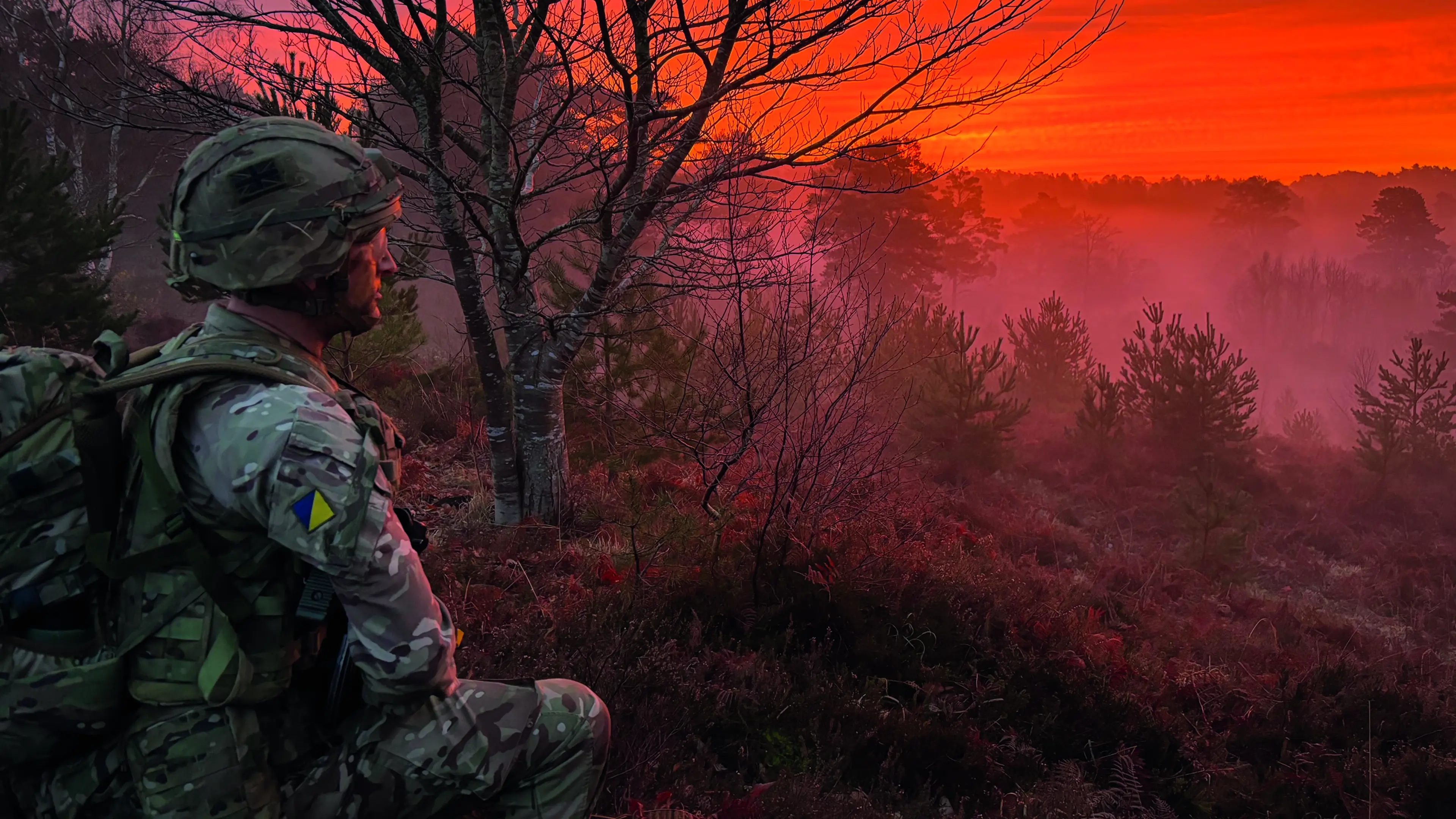 Uniformed soldier crouching and staring into a red sunset over wooded area