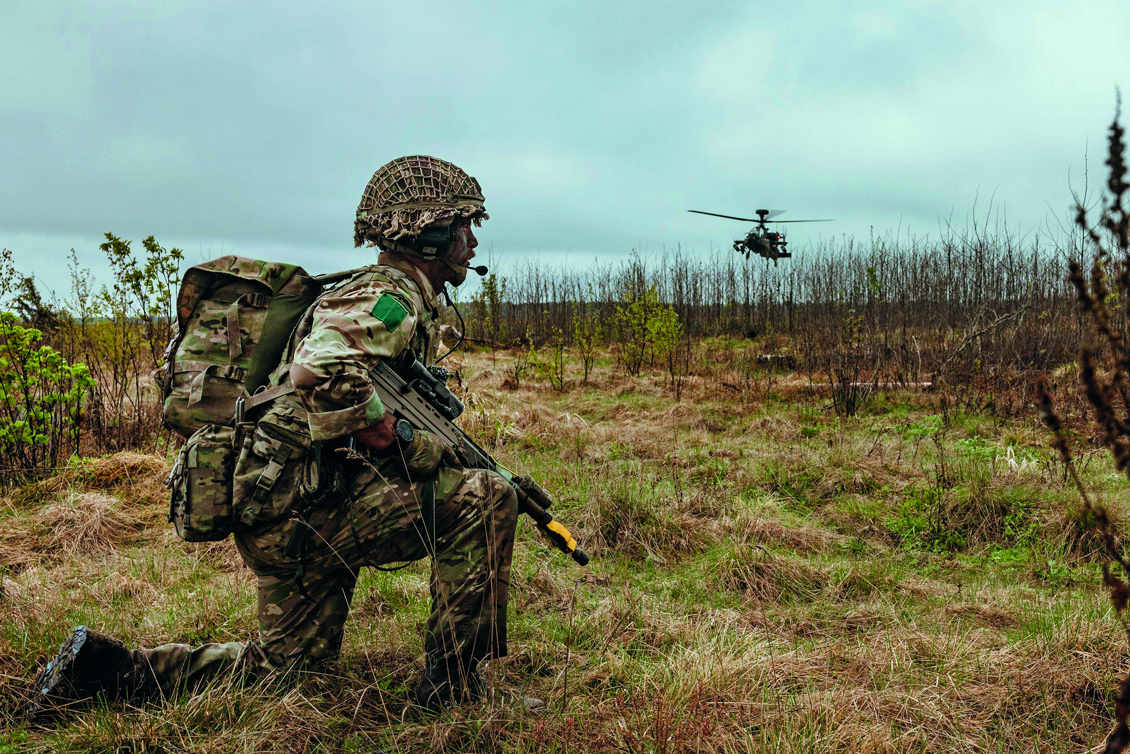 Uniformed soldier crouching in brown grass with helicopter in background