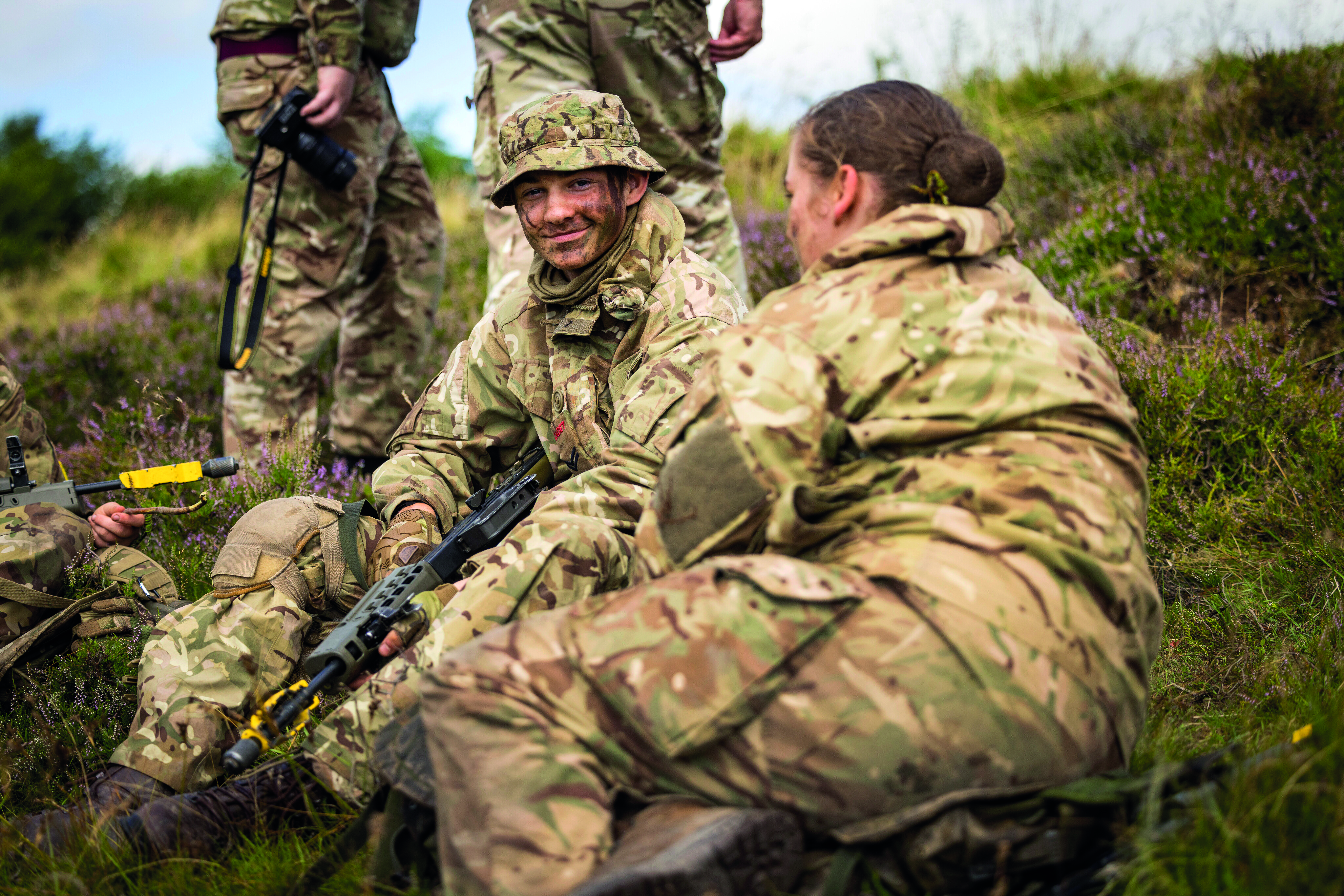 Group of young soldiers sat on grass, focusing on one young man smiling at the camera 
