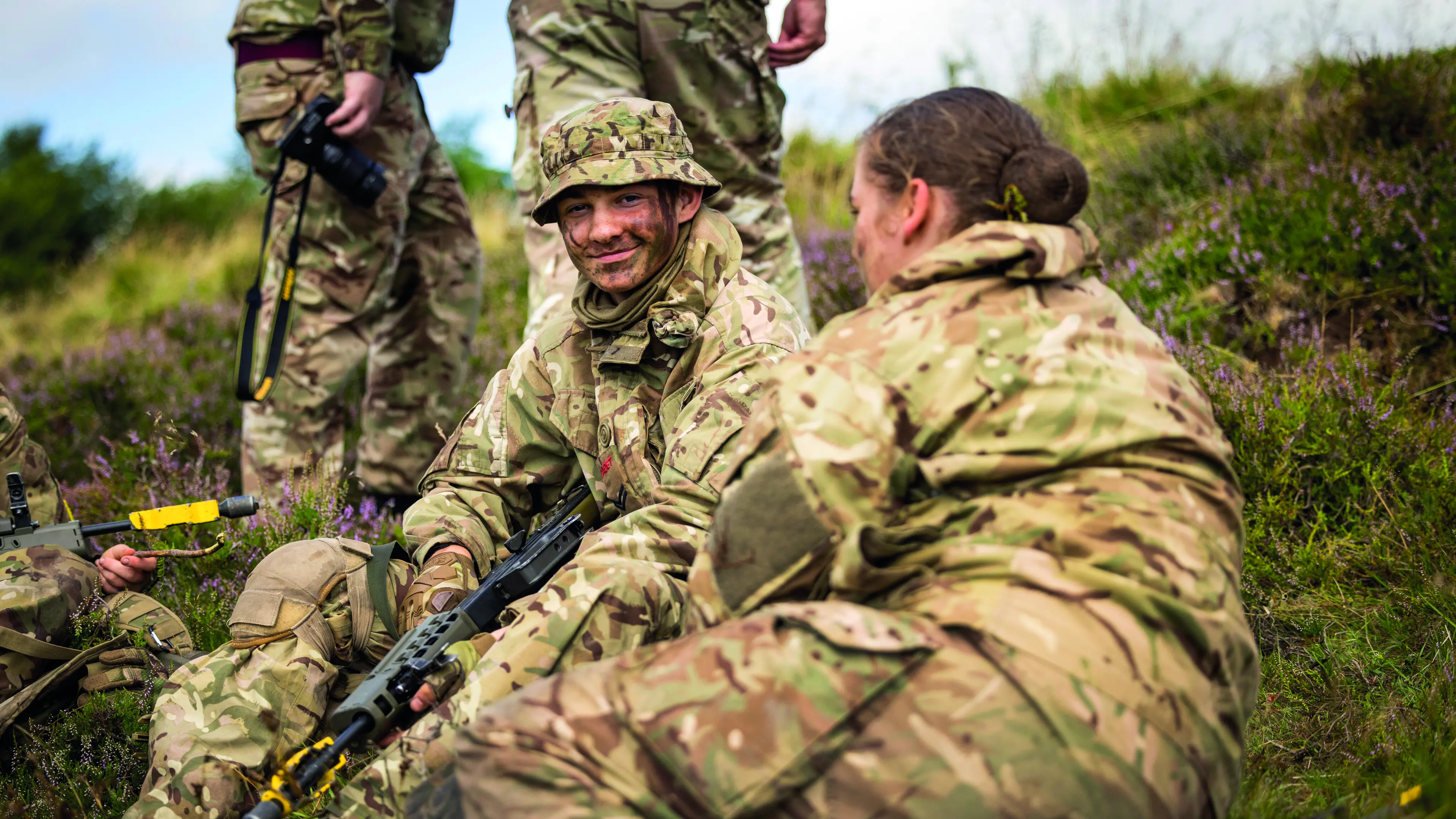 Group of young soldiers sat on grass, focusing on one young man smiling at the camera
