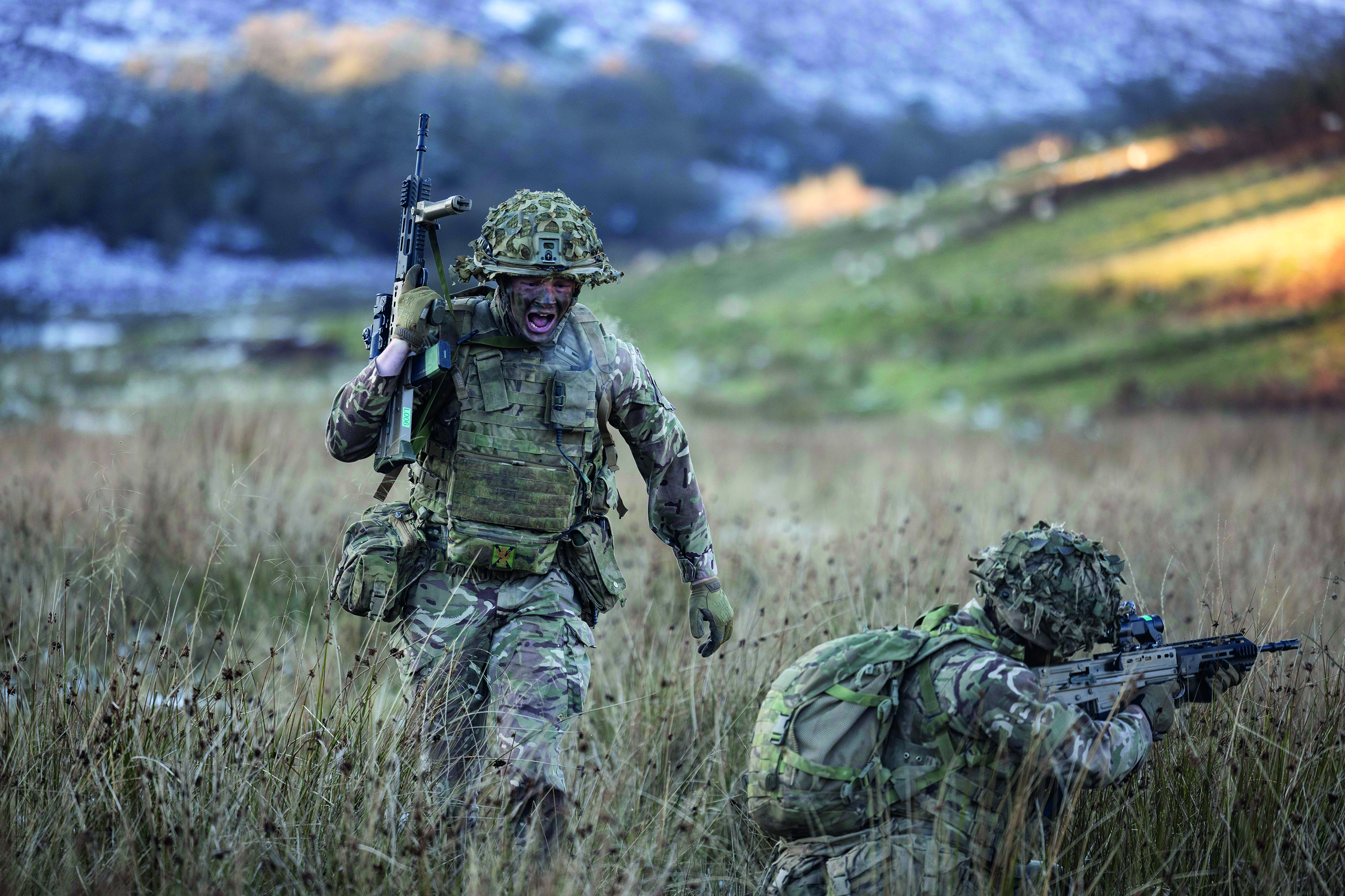 Two soldiers with guns in a grassy field, one is crouched down aiming their gun