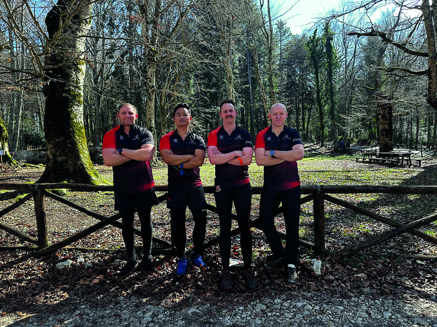 Four men in matching black polos with arms crossed, in front of wooded area