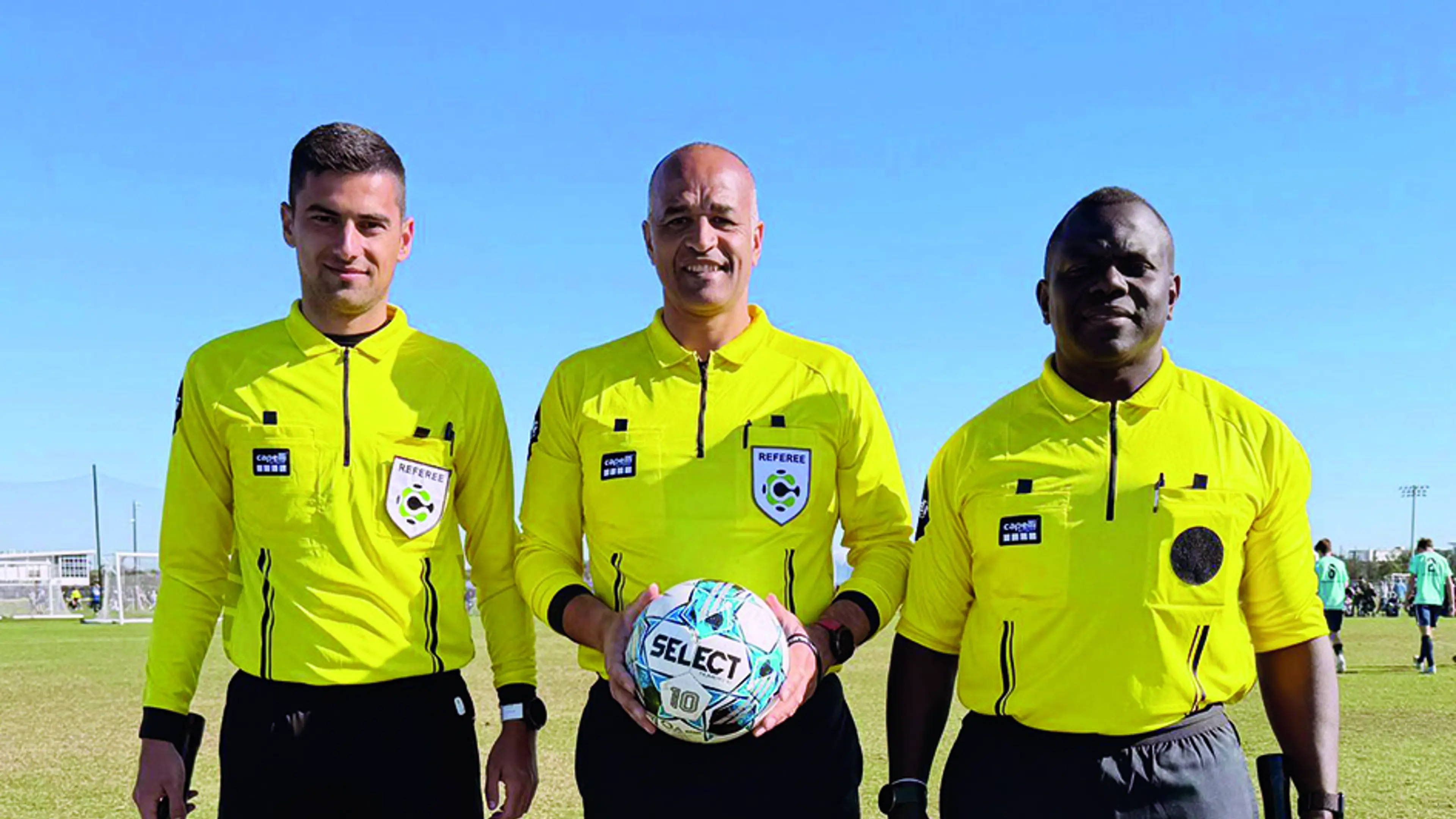 3 men in yellow football shirts smiling at the camera, 1 is holding a football