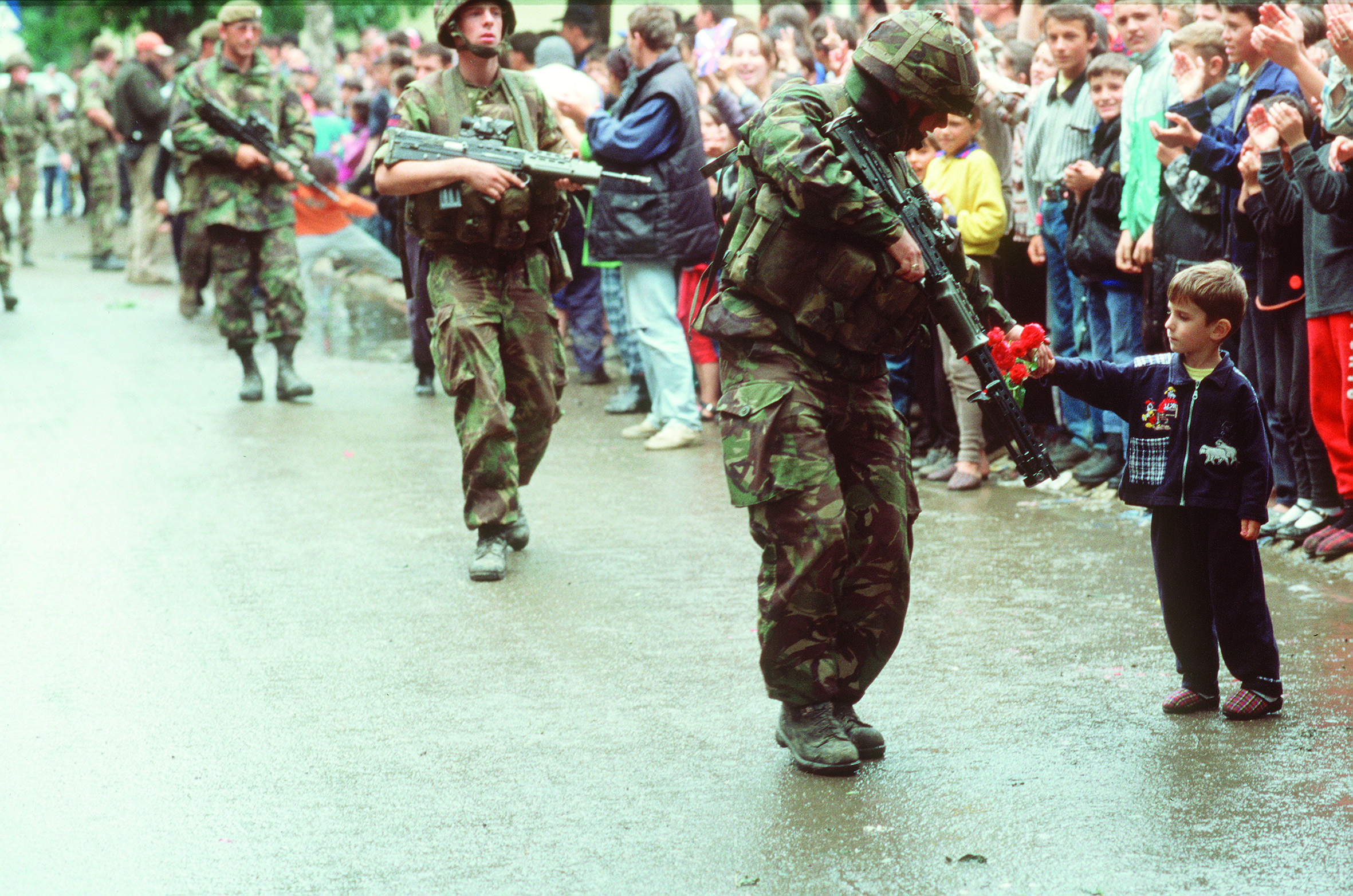 Soldiers with guns walking down a street lined with people, with a child handing roses to the front soldier