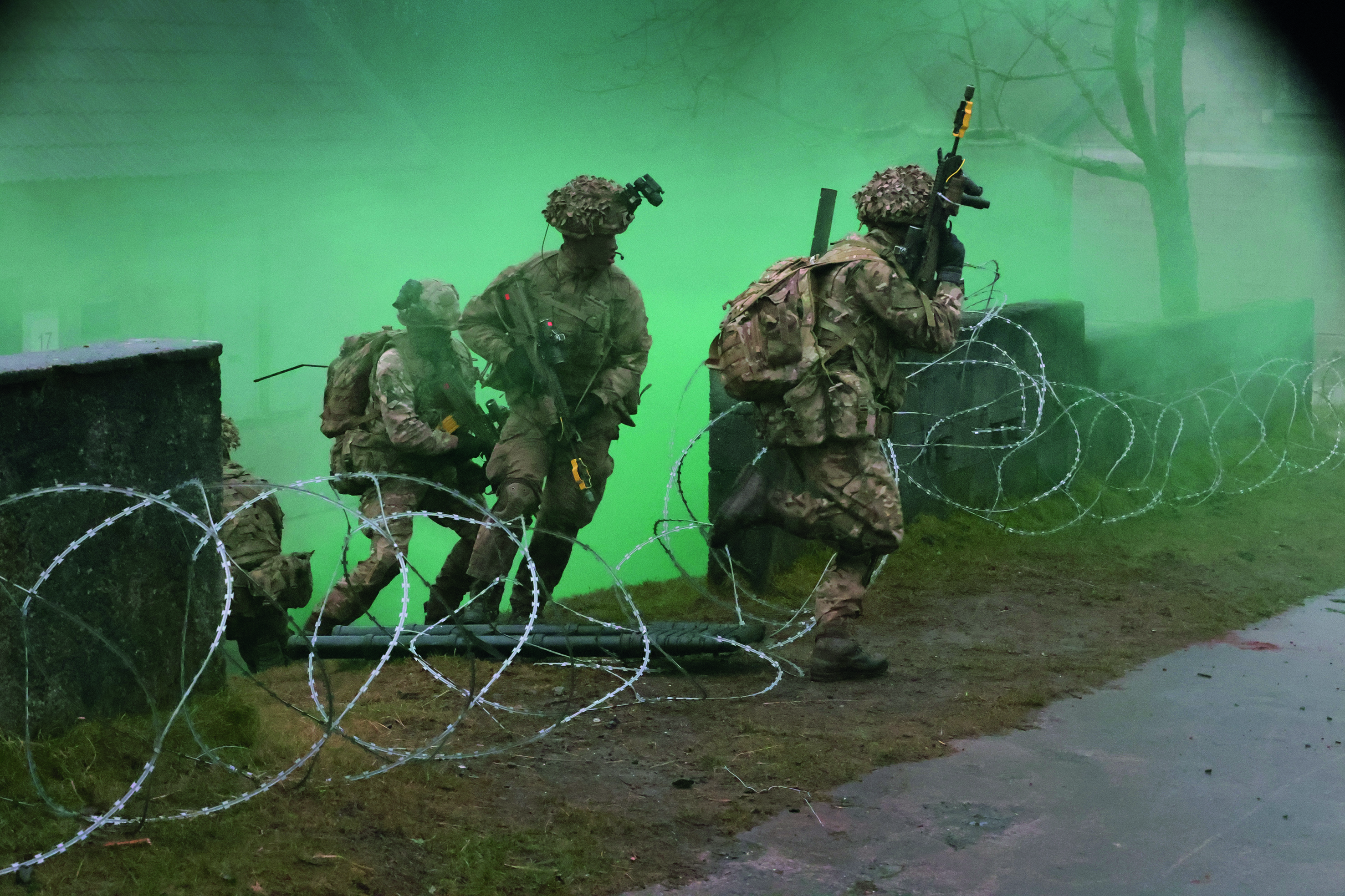 Four soldiers, holding guns and running through barbed wire with green smoke