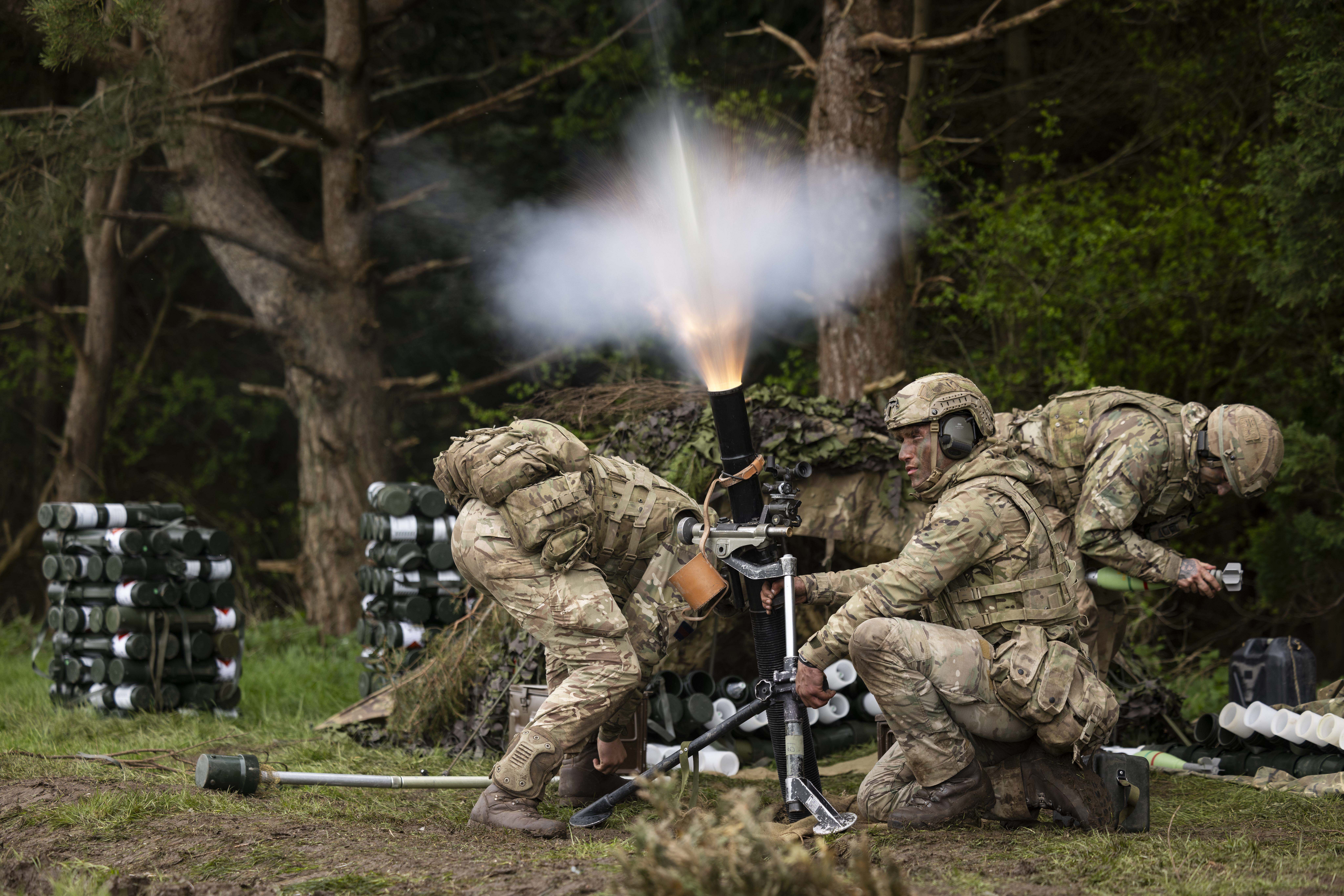 Three uniformed soliders ducking and setting off a cannon device on a bed of grass