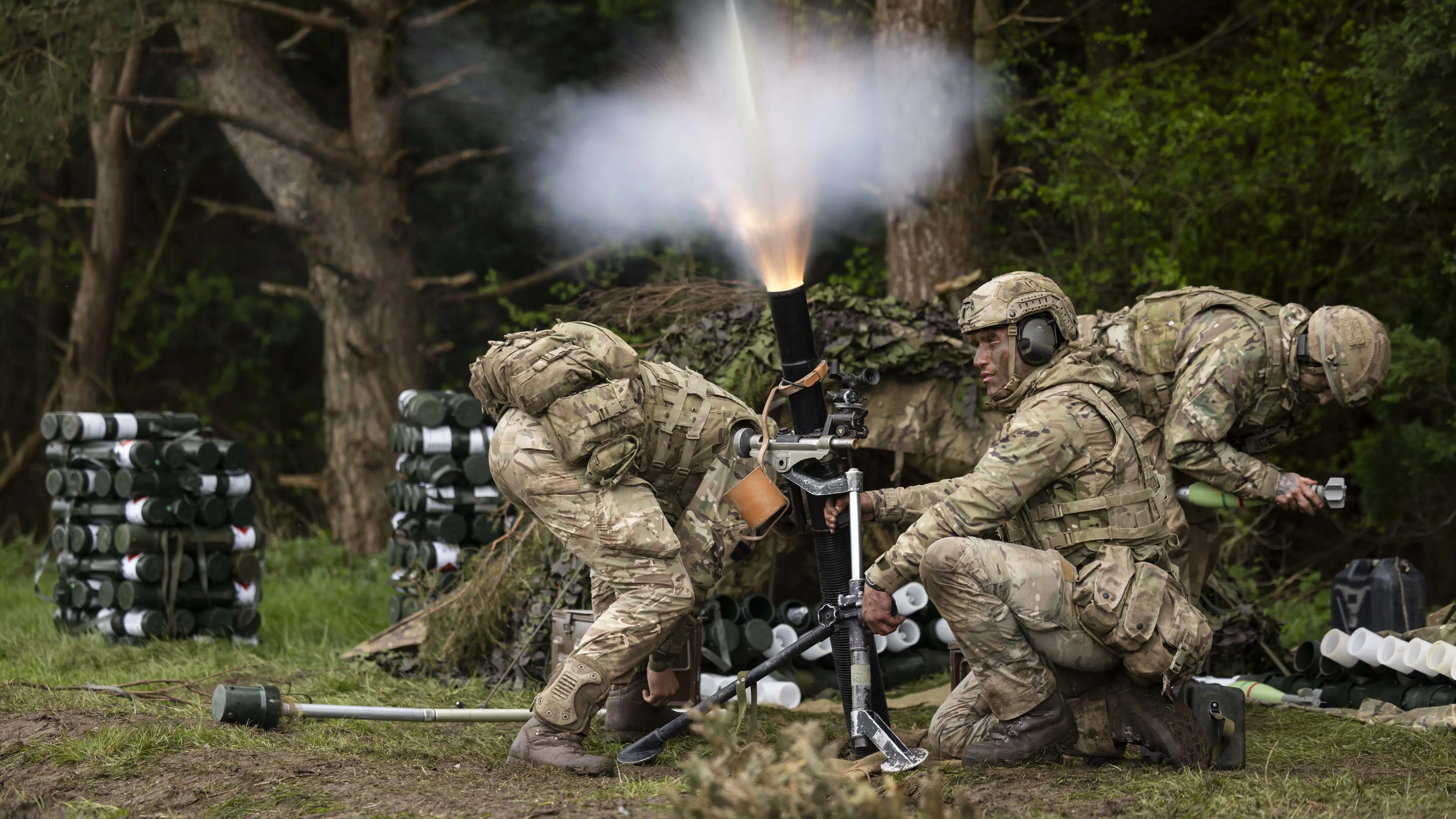 Three uniformed soliders ducking and setting off a cannon device on a bed of grass